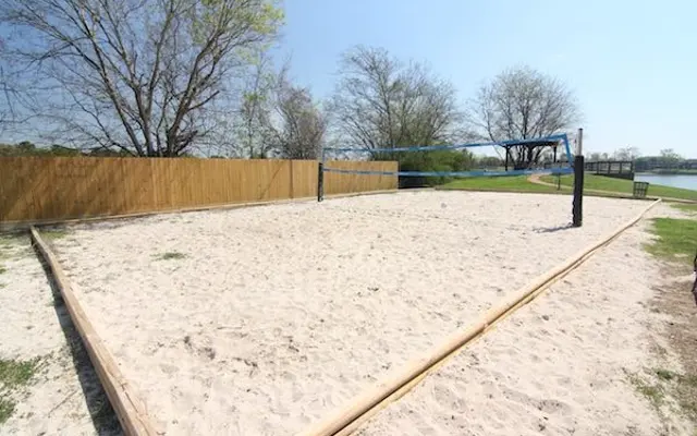 Beach Volleyball Court A sandy beach volleyball court with a net in the center, surrounded by wooden fencing and trees, near a body of water under a clear blue sky.