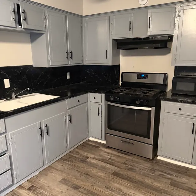 A modern kitchen with gray cabinets and a black countertop, featuring a stainless steel stove, a black microwave, and wooden flooring.