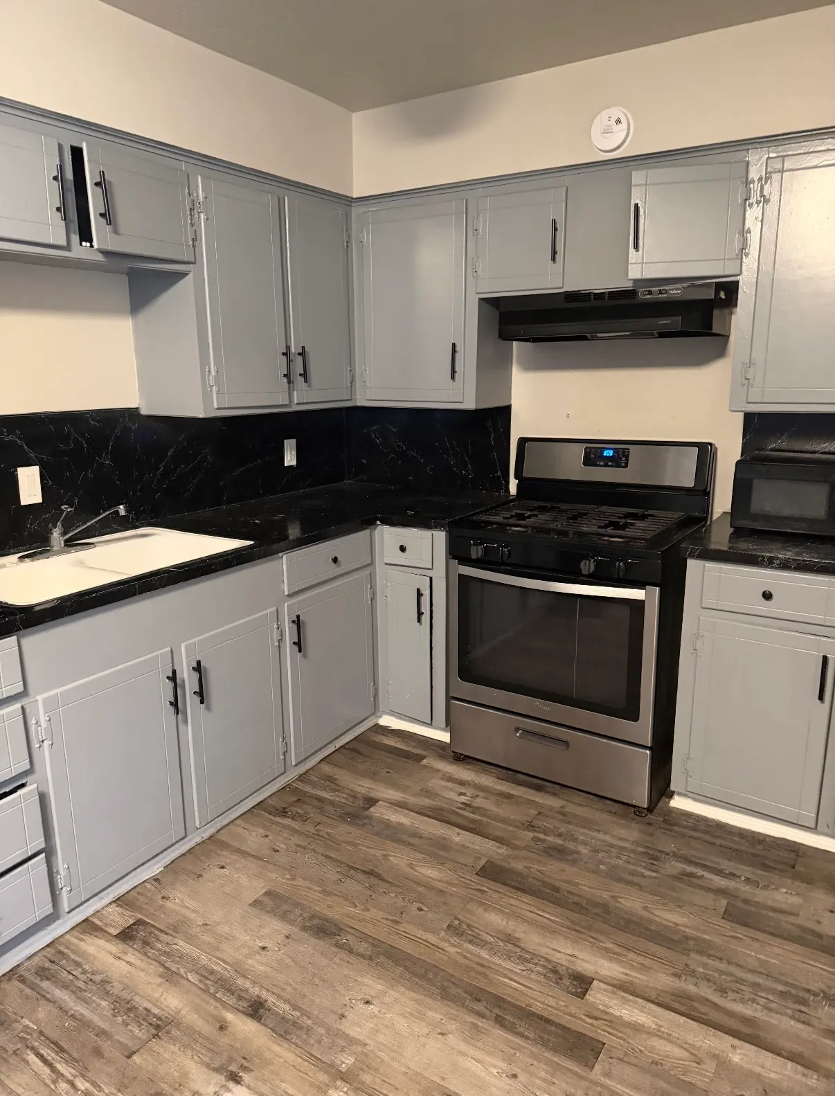 A modern kitchen with gray cabinets and a black countertop, featuring a stainless steel stove, a black microwave, and wooden flooring.