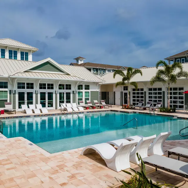 ShoreView A swimming pool with a sun shelf, lounge chairs, umbrellas, and palm trees near a resident clubhouse.