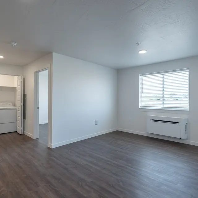 A bright and spacious room featuring a washer and dryer tucked in a closet, with a large window on the right allowing natural light to enter, and a modern air conditioning unit installed underneath.