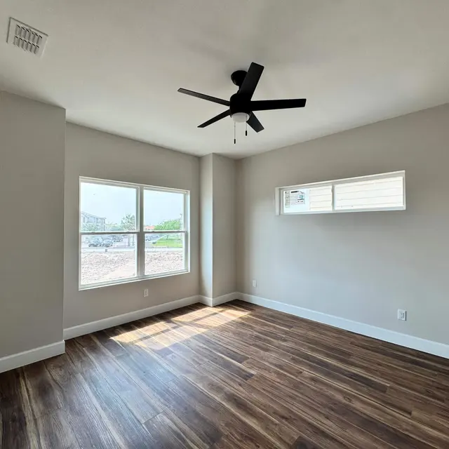 A spacious and bright empty room with wooden flooring, two windows allowing natural light, and a black ceiling fan. The walls are painted in a neutral color.