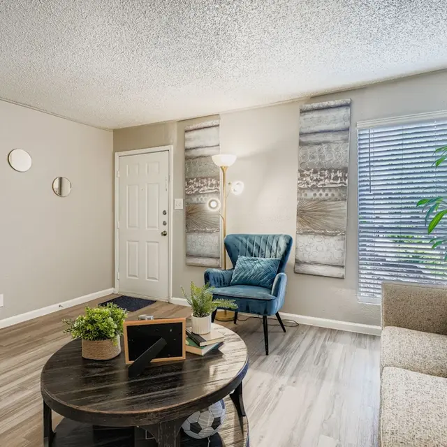 Cozy Living Room Interior A cozy living room featuring a circular coffee table, a blue armchair, and a light-colored sofa with decorative cushions. There are small round mirrors on the wall, and a potted plant next to a window with blinds.