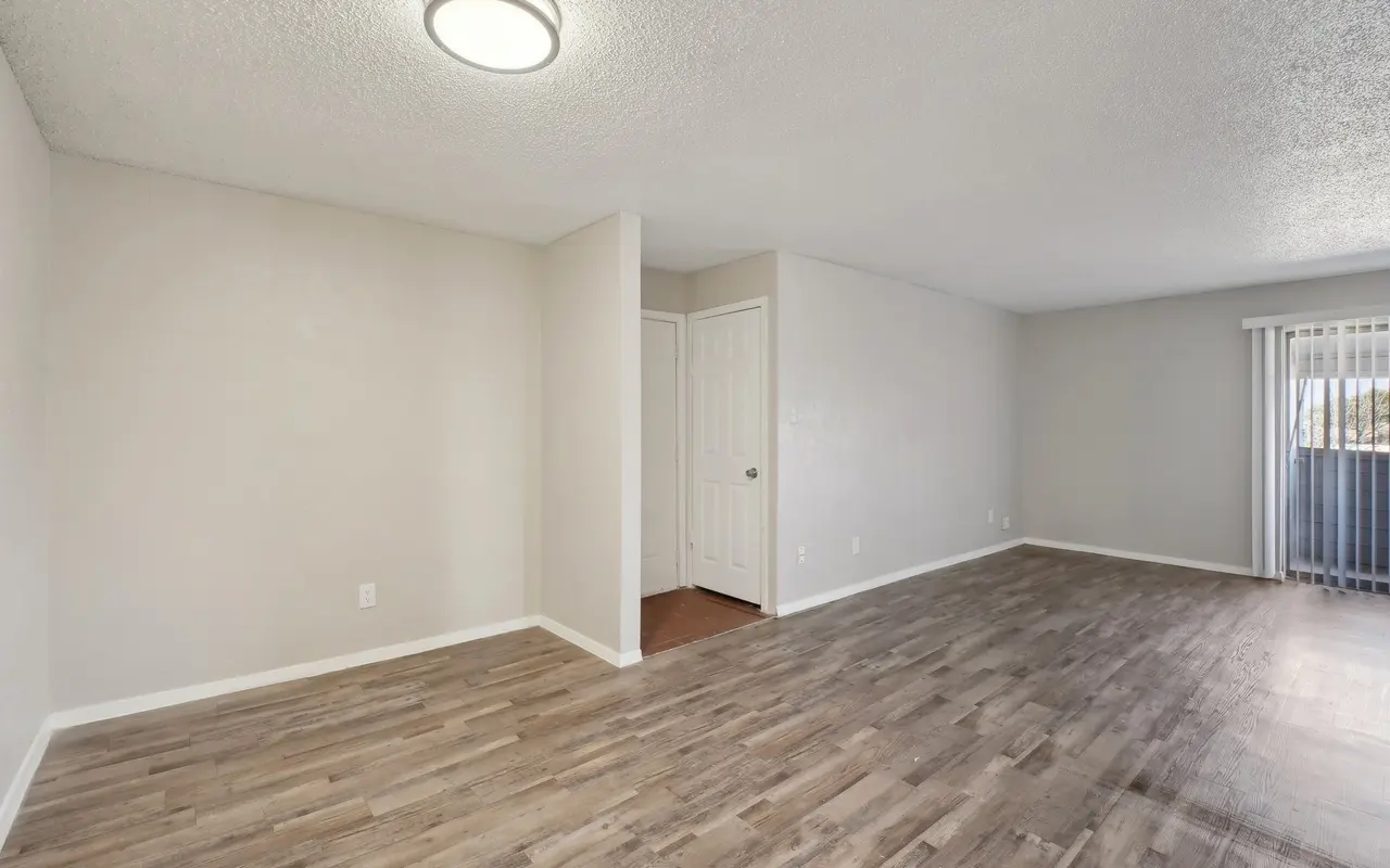 An empty room with light-colored walls and wooden floor. A ceiling light provides overhead illumination. In the background, there is a door leading to another room and a window with vertical blinds.