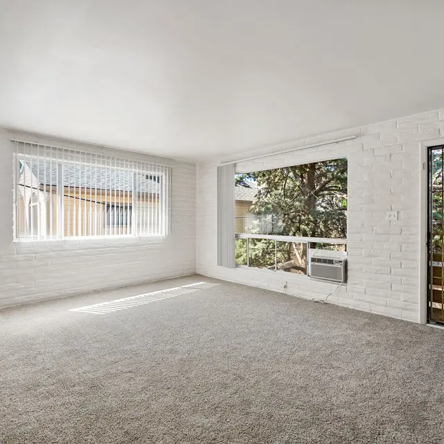A spacious living room with white brick walls and large windows letting in natural light, featuring carpeting in a neutral tone and a sliding glass door leading outside.