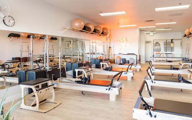 A modern Pilates studio featuring several reformer machines, exercise balls, and large mirrors along the walls, with bright lighting and wooden flooring.