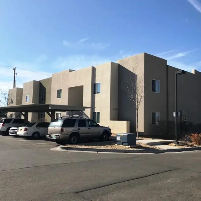 A modern multi-unit apartment building with a flat roof, surrounded by an asphalt parking lot. There are several vehicles parked in front, and a few sparse trees in the landscaping.