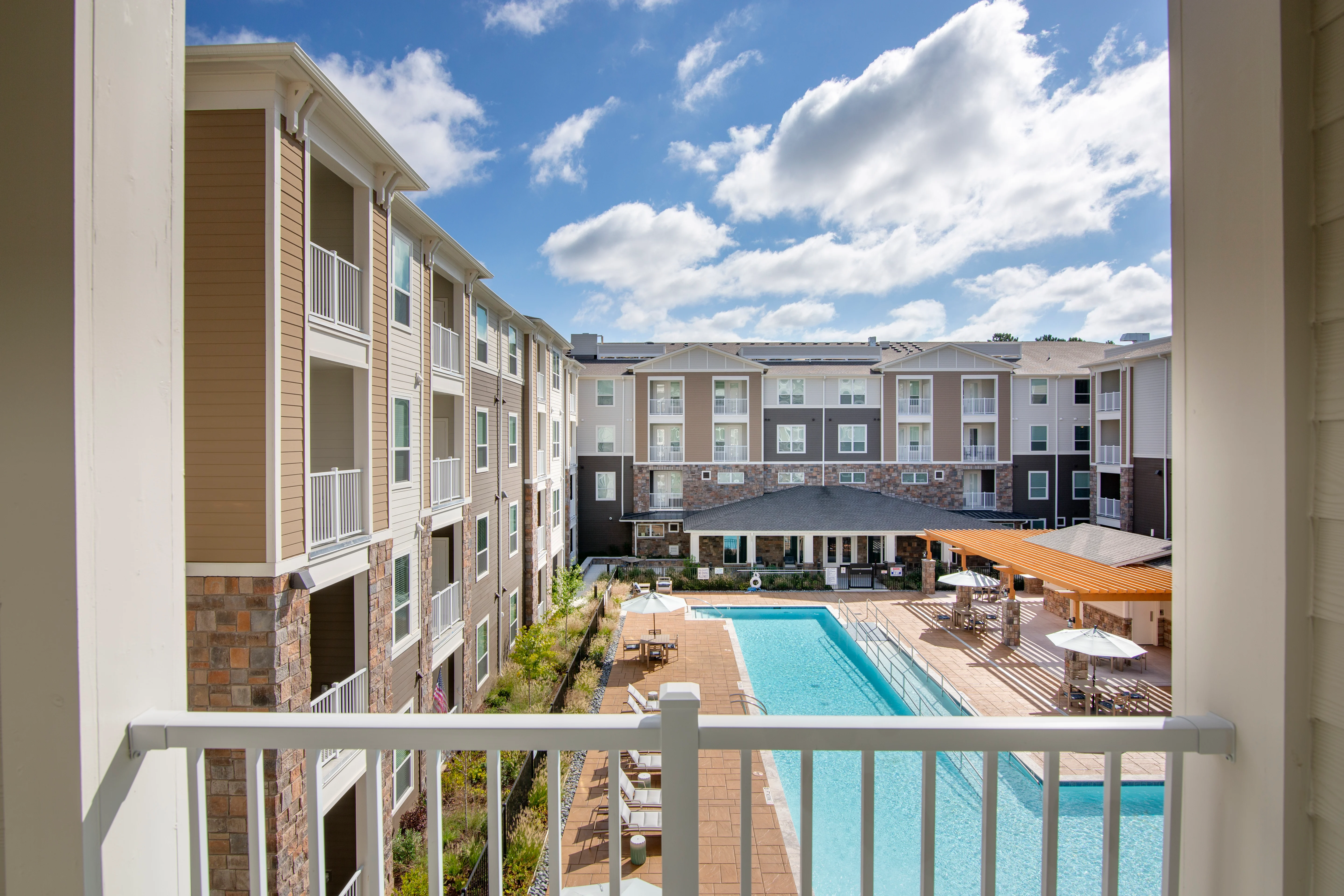 View from a balcony overlooking a modern apartment complex with a swimming pool and lounge area, surrounded by greenery and under a blue sky with scattered clouds.