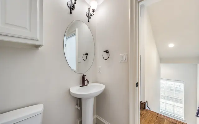 Elegant Modern Bathroom A modern bathroom featuring a small white sink with a mirror above it. The sink has a brown faucet. On the wall beside the sink, there are two wall sconces providing light. The bathroom is painted in light colors, and the door leads to a well-lit hallway. The floor is wooden.