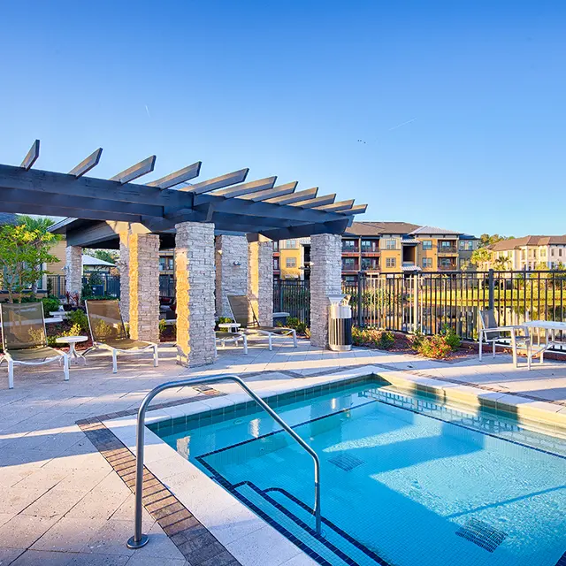 A modern outdoor pool area featuring a small pool with steps, surrounded by lounge chairs and a pergola. The scenery includes green landscaping and buildings in the background under a clear blue sky.