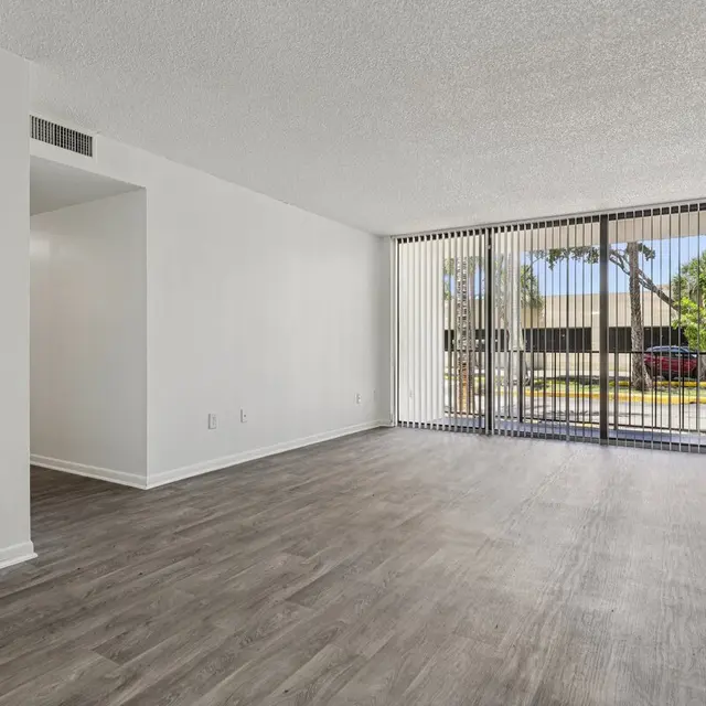 Living Room - Biscayne Apartments A spacious empty living room with a wooden floor and large glass door panels leading outside. The walls are painted white and there are vertical blinds over the door. A small wall-mounted thermostat is visible.