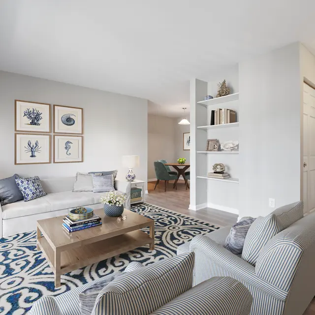 A bright and airy modern living room featuring a light-colored sofa, a wooden coffee table, and decorative artwork on the walls. The room has a cozy rug with a blue pattern, and a small dining area is visible in the background. Shelving units are also present, showcasing decorative items.