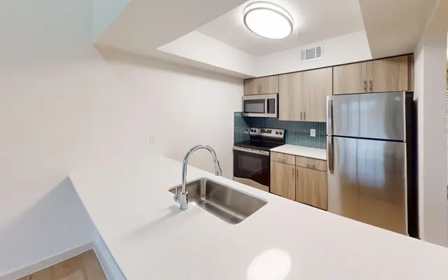 The Gleason A modern kitchen featuring a white countertop, stainless steel appliances, and wooden cabinetry. The sink is centrally located on the island, with a view towards a stove and refrigerator.