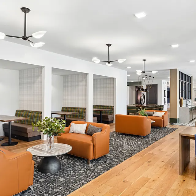A modern lounge area featuring orange leather couches, wooden flooring, and cozy seating booths with plaid upholstery in a well-lit space.