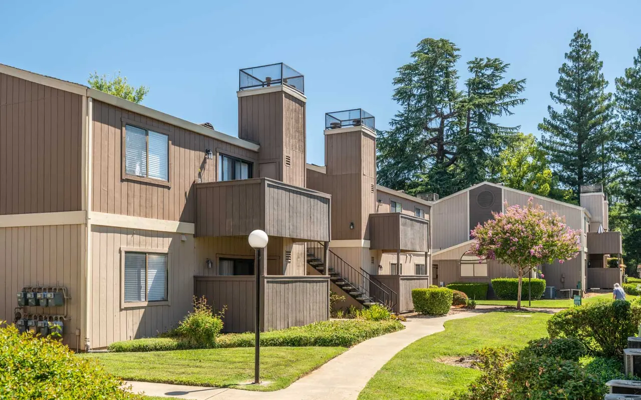 A residential complex with brown wooden buildings and green landscaping, featuring a pathway, lamppost, and trees in the background.