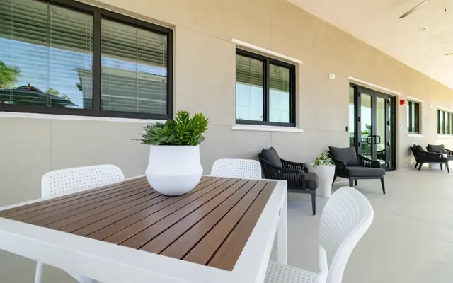 A modern outdoor patio with a white table and brown wooden top, surrounded by white chairs. A succulent plant in a white pot is on the table. In the background, there are large windows and comfortable seating areas.