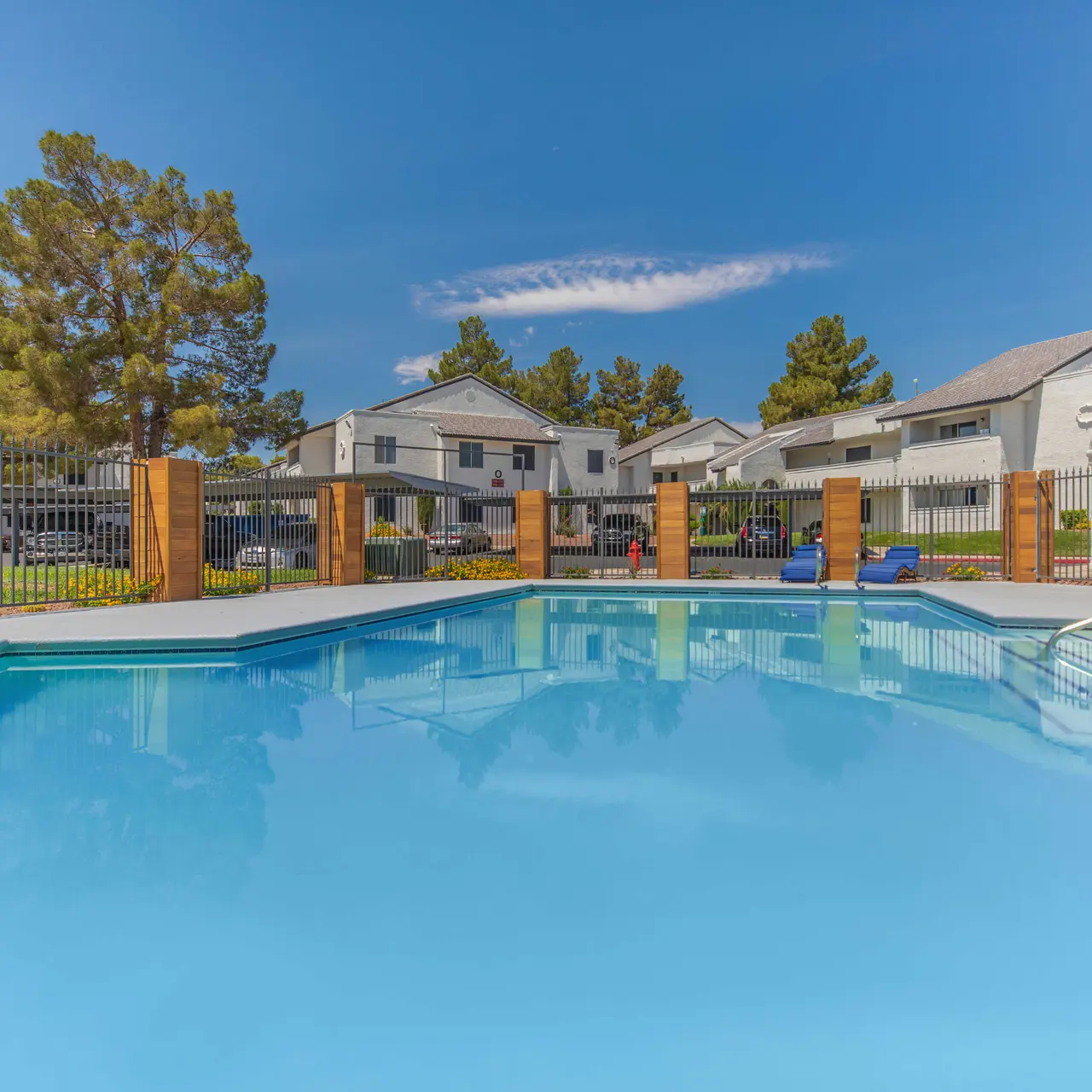A clear blue swimming pool surrounded by wooden fencing, with residential buildings in the background under a bright blue sky.