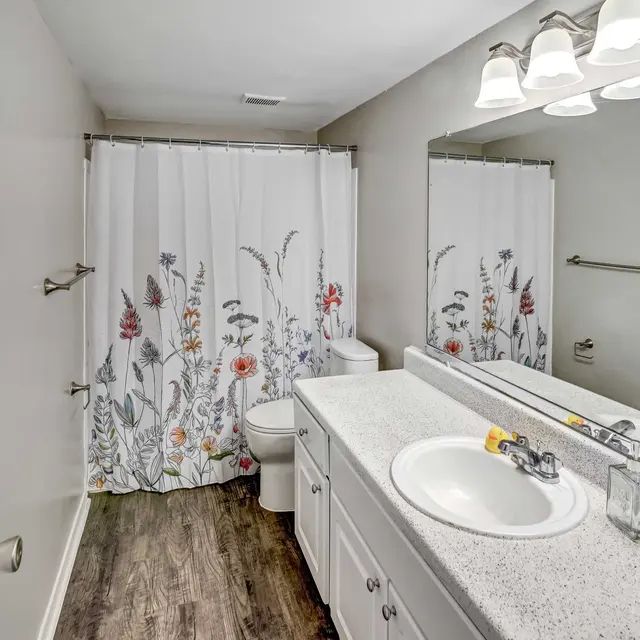 A bright, clean bathroom featuring a floral shower curtain, white vanity with a sink, and modern lighting. The floor is wooden and the walls are painted a soft gray color.