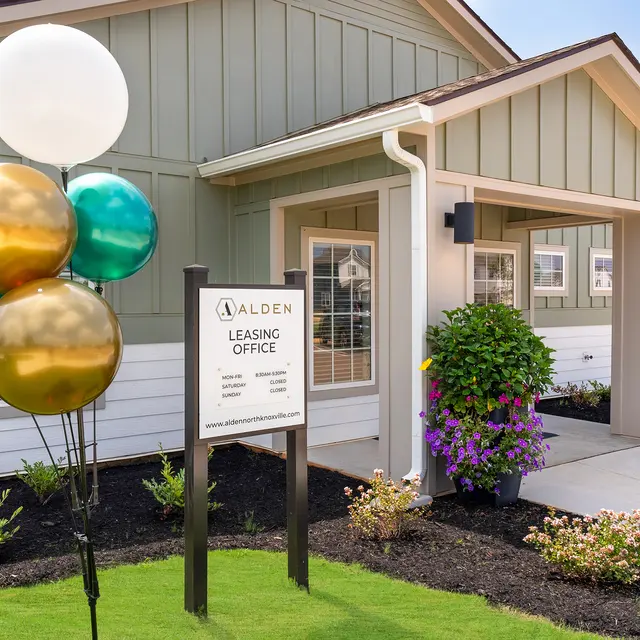 The exterior of a leasing office with green and white walls, featuring a sign that reads 'Alden Leasing Office'. There are balloons in gold, teal, and white, as well as colorful flower pots around the entryway.