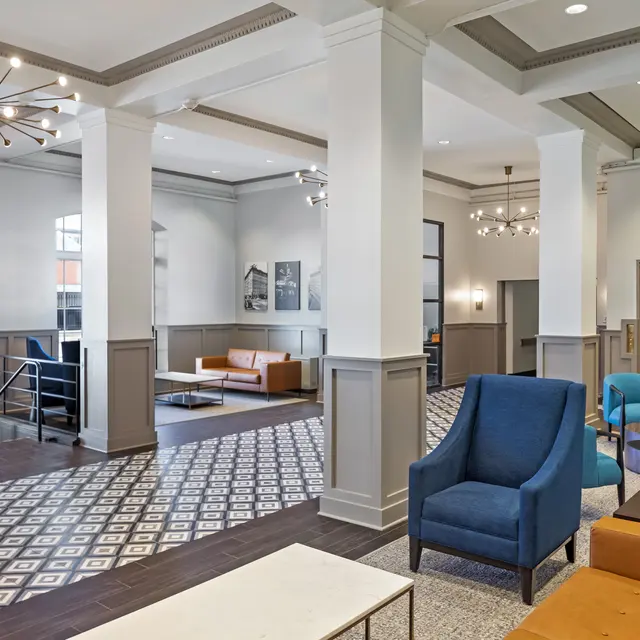 A spacious modern lobby with stylish furniture including blue and orange chairs, a white coffee table, and geometric patterned flooring. Bright lighting and elegant fixtures enhance the atmosphere.