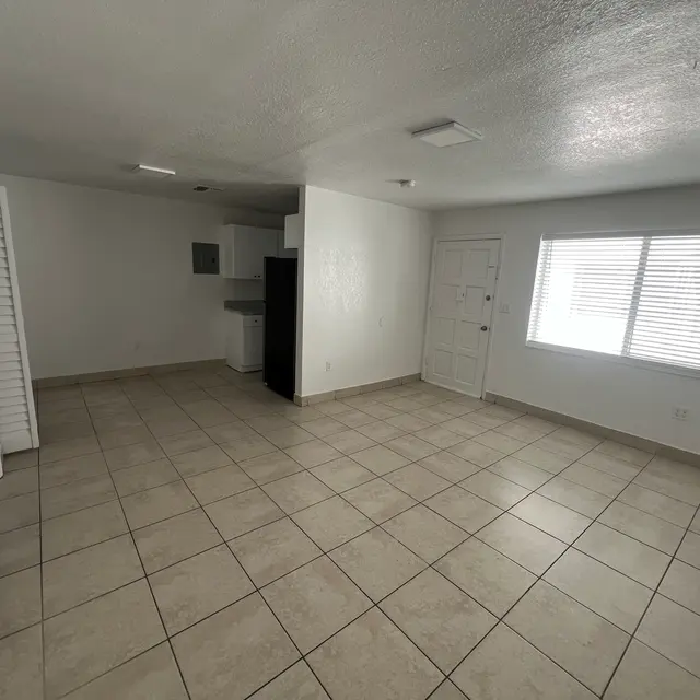 Empty room with light-colored tiled floor, white walls, a kitchen area at the back, and a window with blinds.