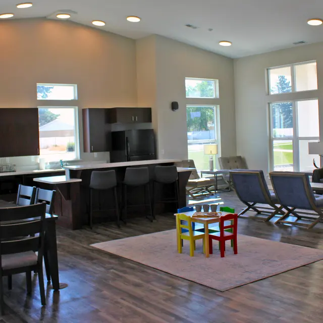 A spacious and modern living area featuring a dining table, kitchen bar stools, seating area with a sofa, coffee table, and a colorful play table for children. Large windows allow natural light to illuminate the room, highlighting the contemporary design.