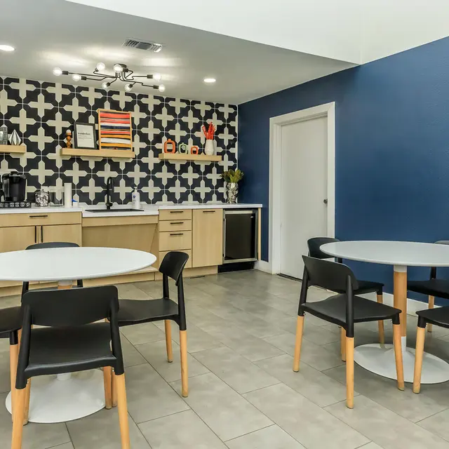 A spacious modern kitchen area featuring two round tables with black chairs, wooden cabinetry, and a patterned wall. A door leads to another room.