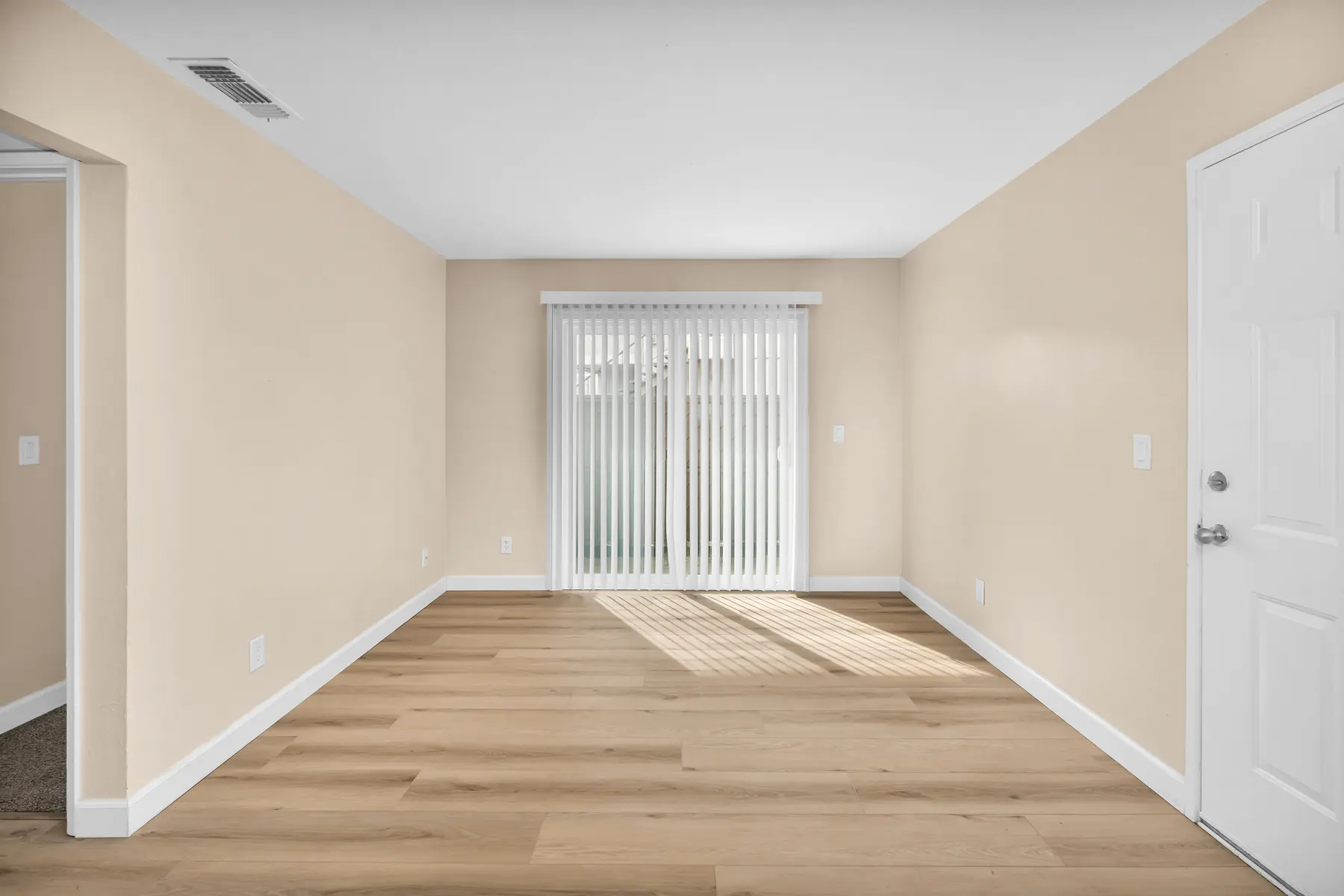 An empty room featuring light-colored walls and wooden flooring, with a sliding glass door covered by vertical blinds letting in natural light.
