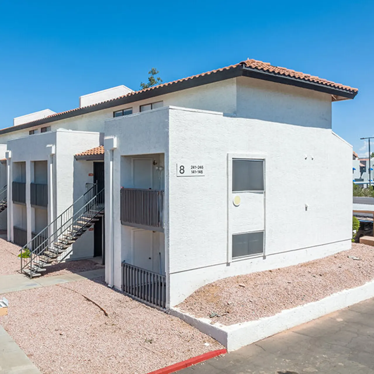 A modern two-story apartment building with a tile roof, featuring external stairs and a minimalist design. The building has a light-colored facade and is surrounded by gravel landscaping.