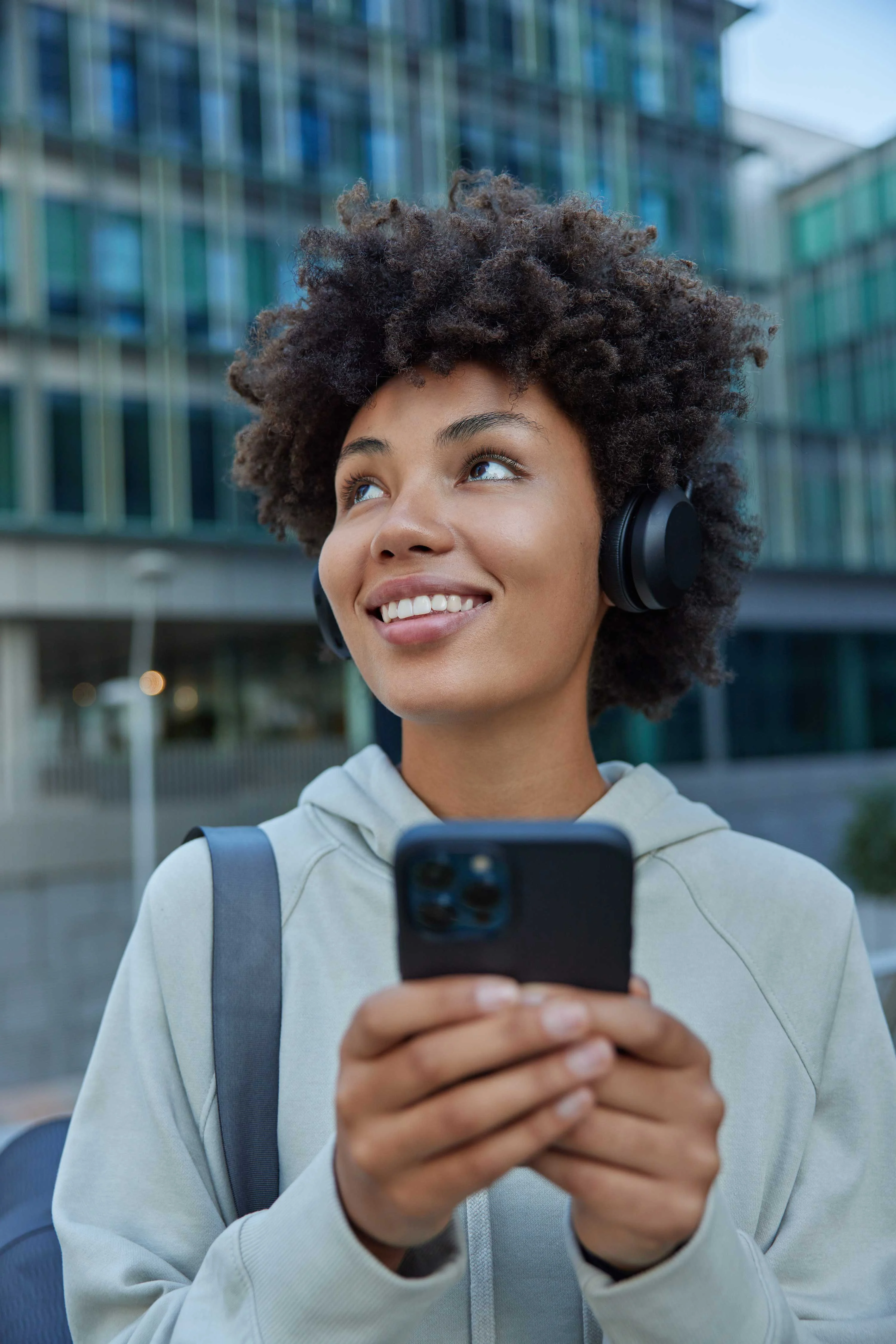 A person with curly hair wearing headphones, smiling while holding a smartphone outdoors in an urban setting.