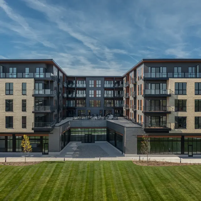 A modern multi-story apartment complex with a symmetrical design, featuring balconies and large windows, surrounded by green lawn space under a blue sky with clouds.
