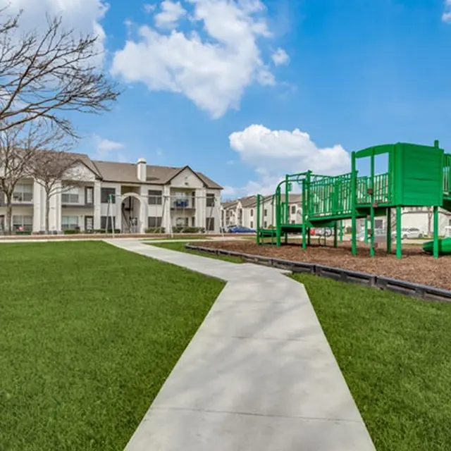 A spacious playground area with a green slide and climbing structure, surrounded by well-maintained lawns and residential buildings.