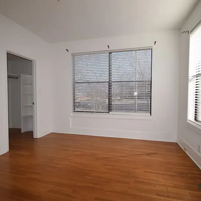 An empty room featuring two large windows with blinds, wooden flooring, and white walls. A closet is visible to the left.