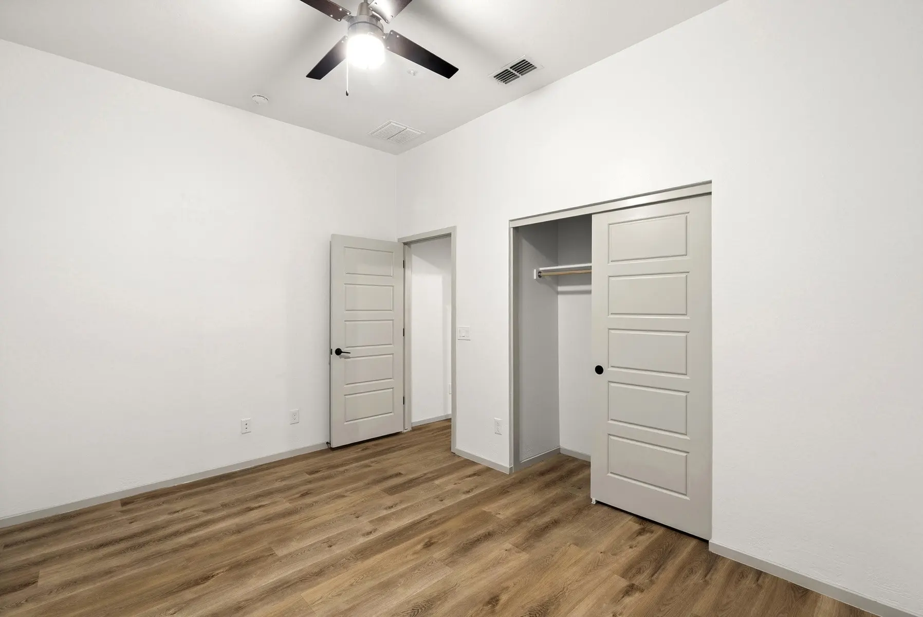 A bright, empty room featuring light wood flooring, a ceiling fan, and two closet doors against white walls.