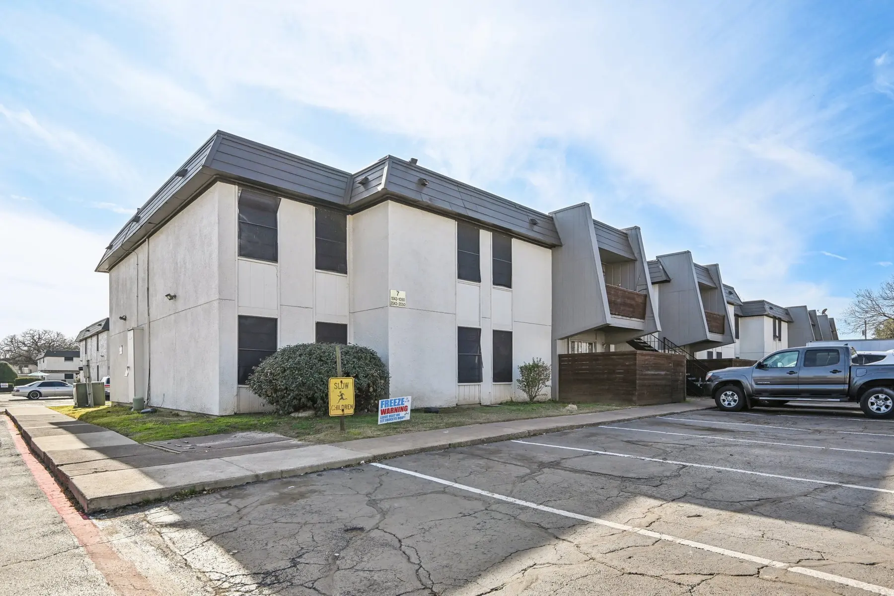 View of an apartment complex with a parking area in the foreground, featuring a two-story building and a few parked vehicles in bright weather.