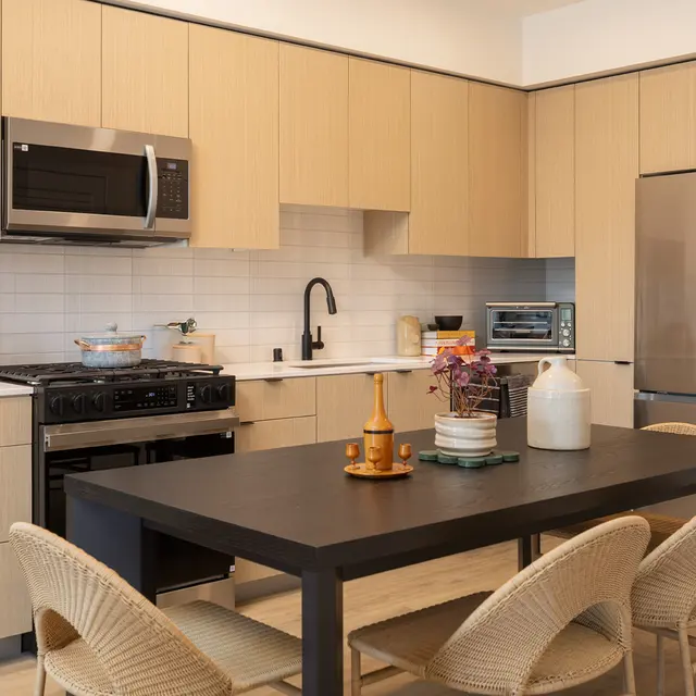 A modern kitchen featuring light wood cabinetry, stainless steel appliances, and a large black dining table surrounded by rattan chairs.