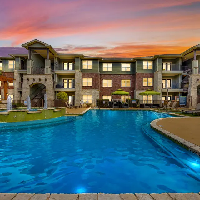 A luxurious swimming pool at an apartment complex during sunset. The pool features a fountain and is surrounded by comfortable lounge chairs. The building has multiple stories with balconies and vibrant decorations.