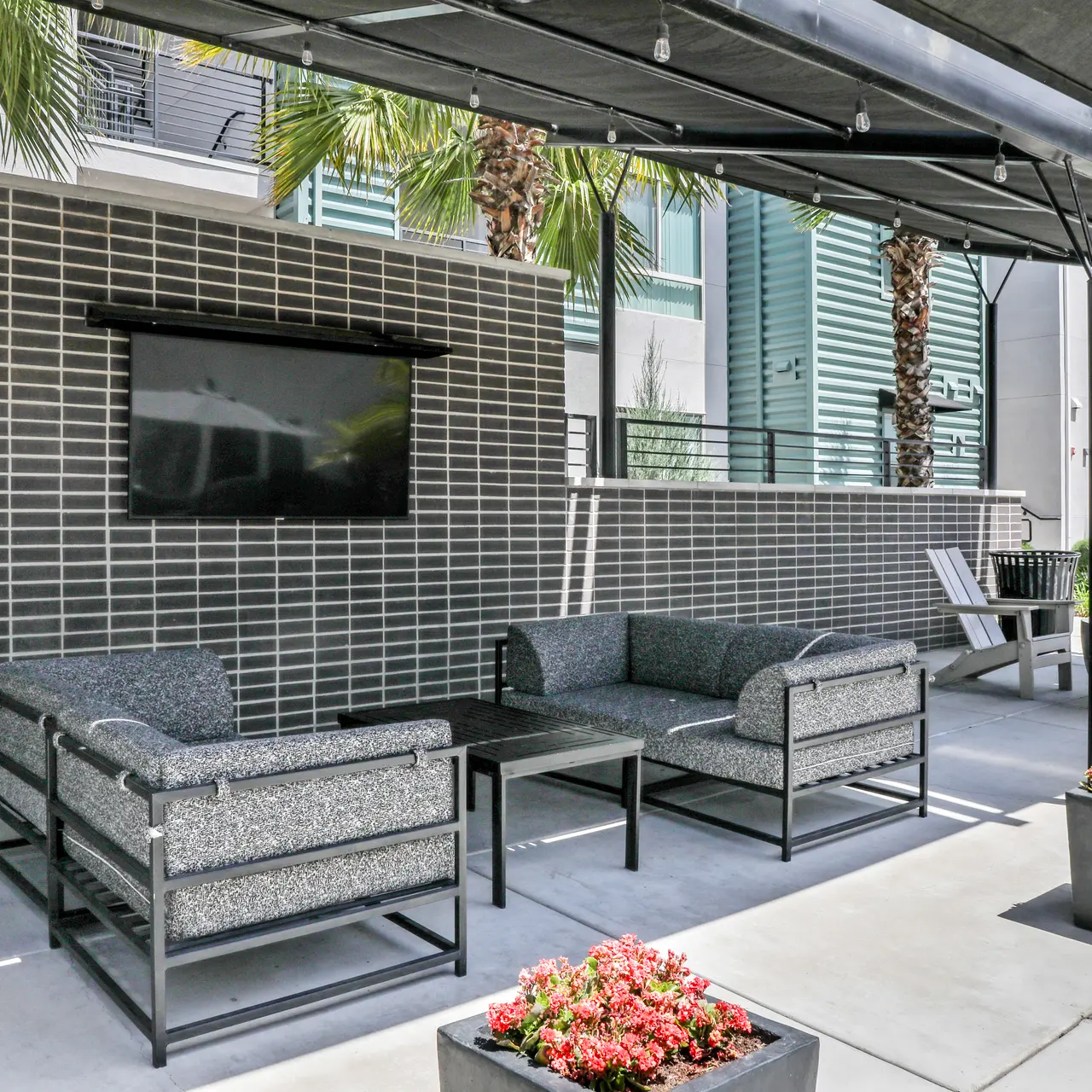 Outdoor lounge area with a seating arrangement and a television mounted on the wall, surrounded by palm trees and planters with flowers.