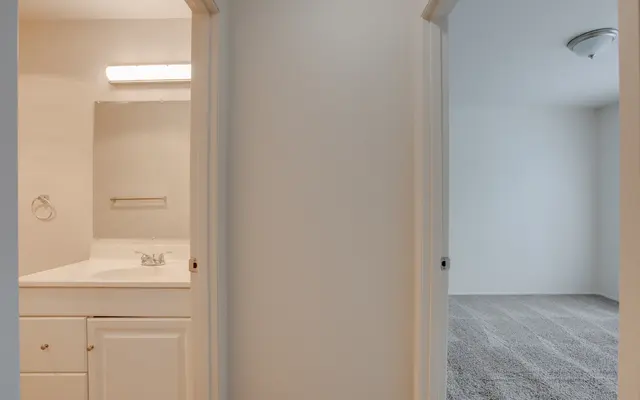 A view from a hallway showing a bathroom on the left with a sink and mirror, and a carpeted bedroom on the right with a light-colored wall.