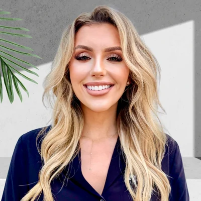 A smiling woman with long blonde hair, wearing a dark blouse, stands against a light-colored wall with a green plant in the background.