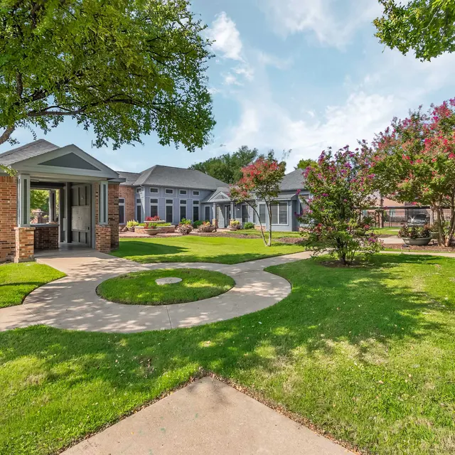 A serene garden area featuring paved paths, green lawns, trees, and flowering plants in a residential setting.
