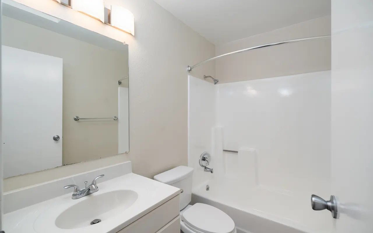 A clean and modern bathroom featuring a white bathtub with a curved shower curtain rod, a single sink vanity with a large mirror, and a toilet.