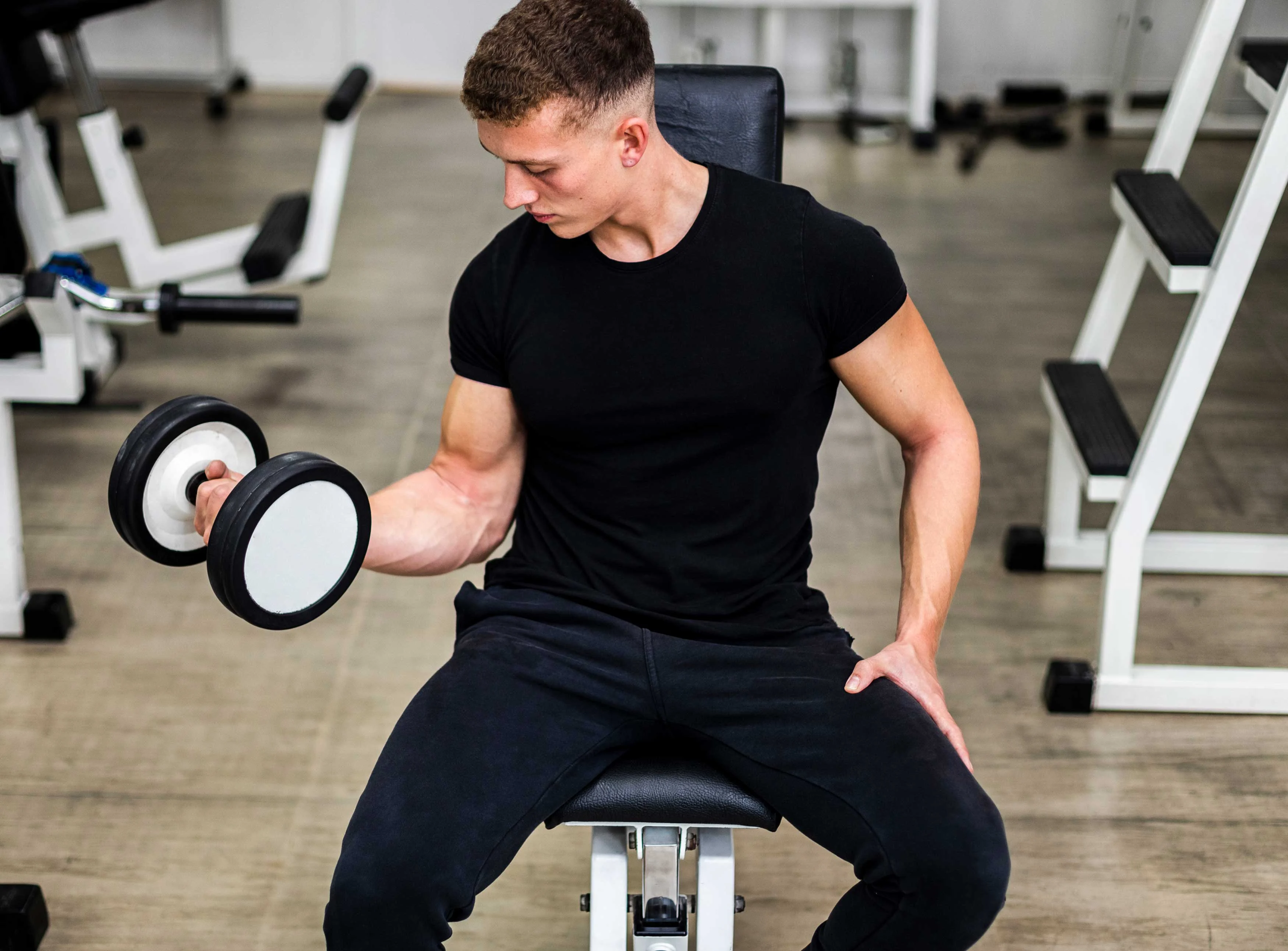 A young man with a muscular build sitting on a bench in a gym, lifting a dumbbell with one arm while looking focused.