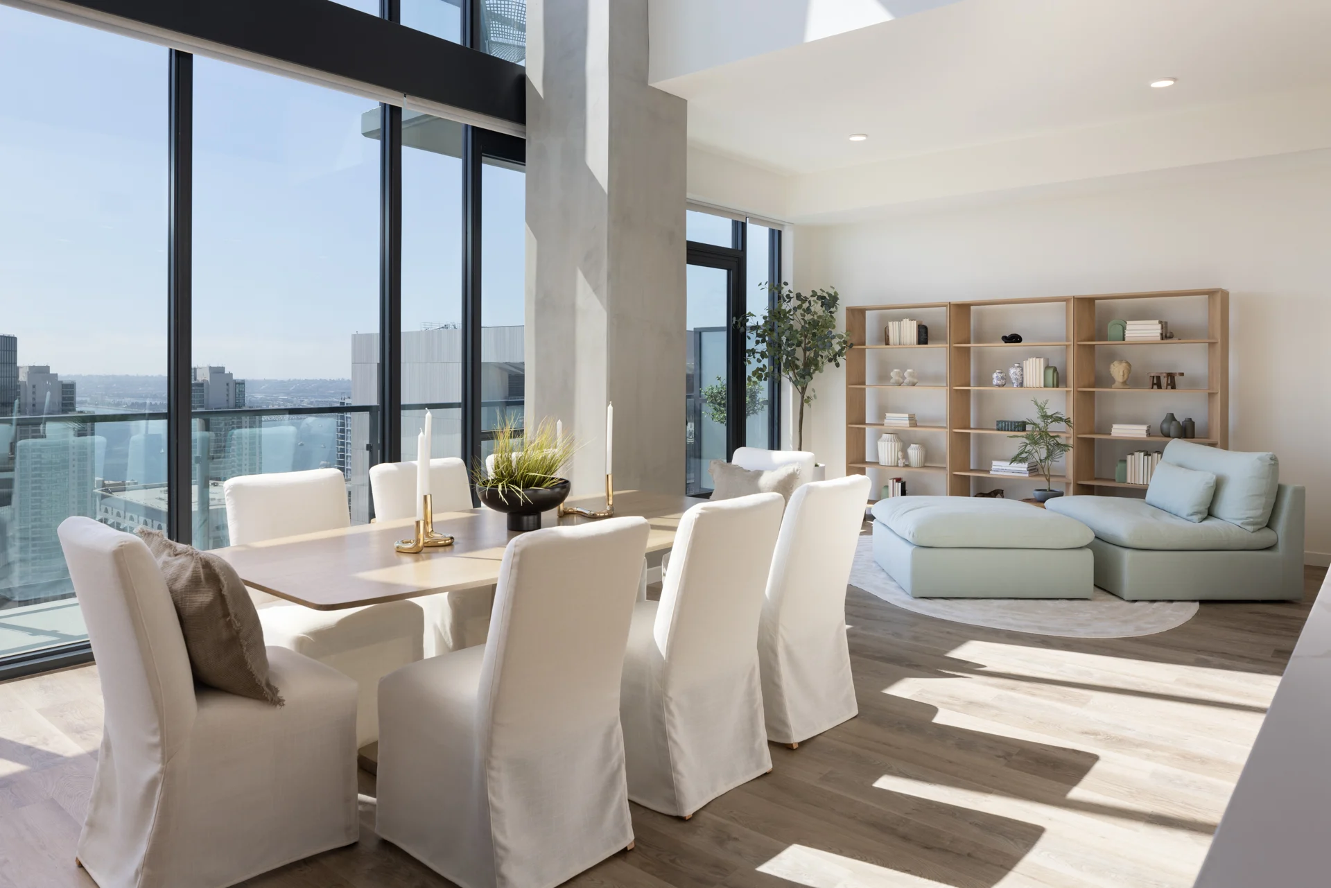 A modern dining and living area featuring a table with white chairs, large windows displaying a cityscape, and a cozy seating area with light-colored furniture.