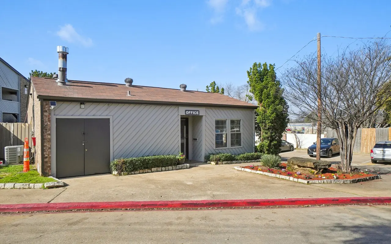 A small office building with a gray exterior featuring a sign that reads 'OFFICE.' The building has a chimney, windows, and is surrounded by neat landscaping. A pathway leads to the entrance, and vehicles are parked nearby.