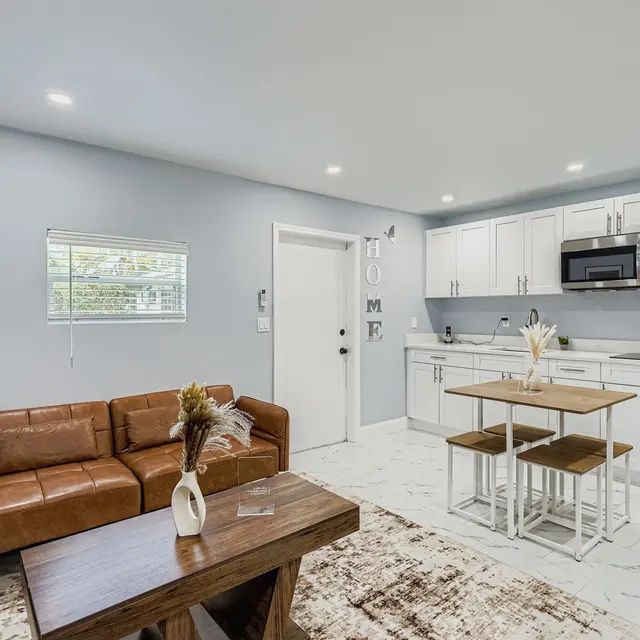 A stylish and modern living space featuring a brown leather sofa, a wooden coffee table, and a dining area with a small table and stools. The kitchen area includes white cabinets, a microwave, and an open layout with ample natural light from the windows.