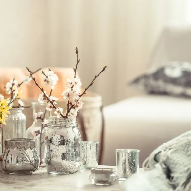 A cozy setting with blooming branches in jars, decorative candles, and a soft blanket on a marble table, with a blurred background.