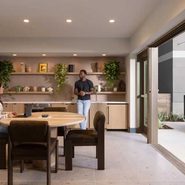 A modern meeting space featuring a round wooden dining table with six chairs, two people engaged in conversation, surrounded by shelves decorated with plants and art. A large glass door opens to an outdoor area with a seating arrangement.
