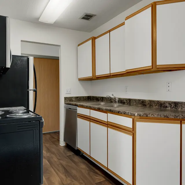Regency Park Apartments Regency Park - A modern kitchen featuring black appliances including a stove and dishwasher, white cabinetry with wooden accents, and a granite countertop.