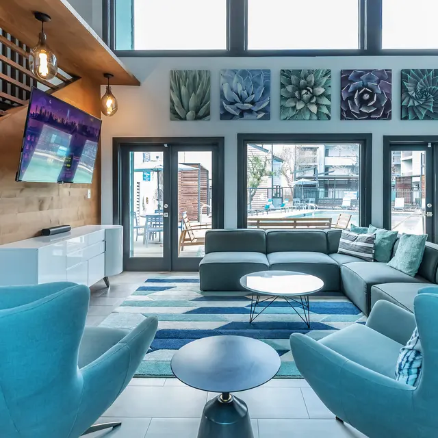 A modern living room featuring a light-colored sectional sofa, a round coffee table, and teal armchairs. The walls are adorned with floral art, and large windows provide natural light. A television is mounted on the wall, and there is a staircase leading to an upper level.